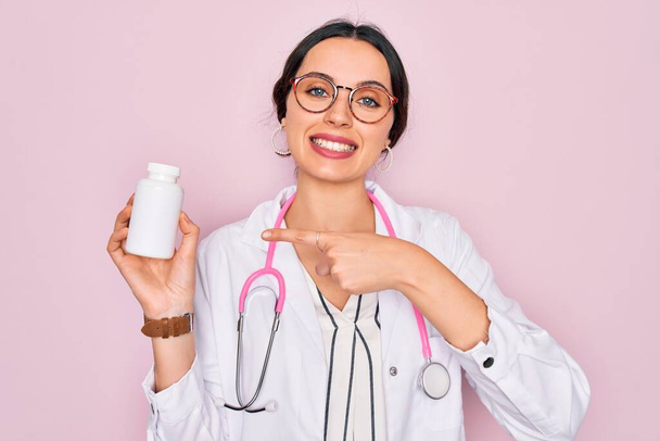 Young beautiful doctor woman with blue eyes wearing stethoscope holding medicine pills very happy pointing with hand and finger - Photo, Image
