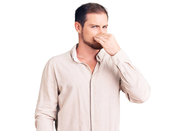 Νεαρός όμορφος καυκάσιος άντρας που φοράει καθημερινά ρούχα μυρίζοντας κάτι βρωμερό και αηδιαστικό, ανυπόφορη μυρωδιά, κρατώντας την αναπνοή του με δάχτυλα στη μύτη. άσχημη μυρωδιά  - Φωτογραφία, εικόνα