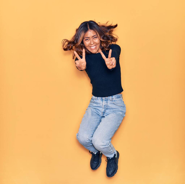 Jonge mooie latijnse vrouw die blij lacht. Springen met een glimlach op het gezicht doen overwinning teken over geïsoleerde gele achtergrond - Foto, afbeelding