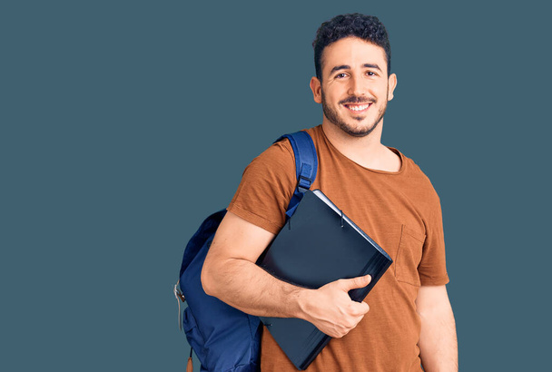Joven hombre hispano usando mochila de estudiante sosteniendo aglutinante mirando positiva y feliz de pie y sonriendo con una sonrisa confiada mostrando los dientes
  - Foto, Imagen