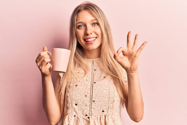 Young beautiful blonde woman holding coffee doing ok sign with fingers, smiling friendly gesturing excellent symbol  - Фото, зображення