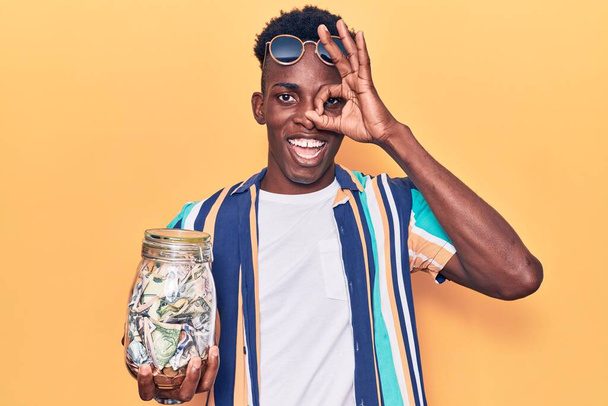 Νεαρός Αφρικάνος Αμερικανός άνδρας κρατώντας βάζο με αποταμιεύσεις χαμογελώντας χαρούμενος κάνει εντάξει σημάδι με το χέρι στο μάτι κοιτάζοντας μέσα από τα δάχτυλα  - Φωτογραφία, εικόνα