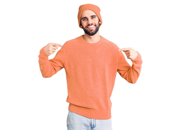 Jonge knappe man met baard met casual trui en wollen pet die er zelfverzekerd uitziet met een glimlach op het gezicht, zich wijzend met vingers trots en gelukkig.  - Foto, afbeelding
