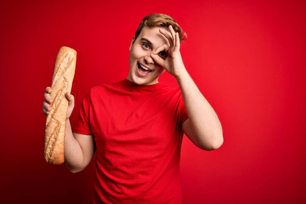 jovem bonito ruiva homem segurando fresco caseiro pão sobre isolado vermelho fundo sorrindo feliz fazendo ok sinal com mão no olho olhando através de dedos
 - Foto, Imagem
