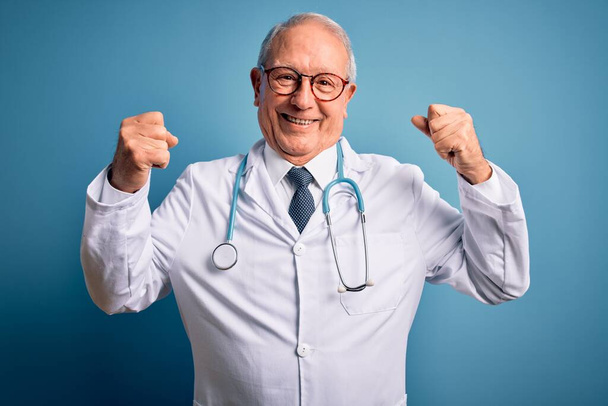 Hombre médico de pelo gris senior con estetoscopio y abrigo médico sobre fondo azul que muestra los músculos de los brazos sonriendo orgulloso. Concepto Fitness
. - Foto, imagen