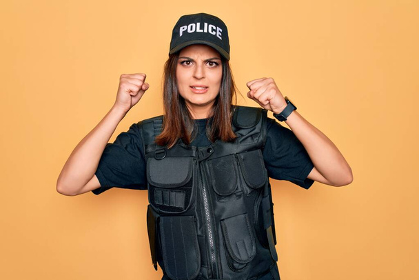 Молодая красивая брюнетка-полицейский в полицейской форме пуленепробиваемая и шапочка злая и безумная поднимающая кулаки разочарованная и разъяренная, крича от злости. Ярость и агрессивность. - Фото, изображение