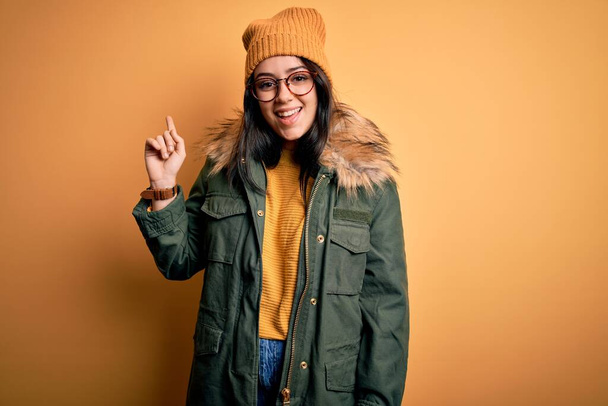 Young brunette woman wearing glasses and winter coat with hat over yellow isolated background with a big smile on face, pointing with hand and finger to the side looking at the camera. - Фото, зображення