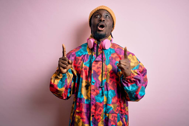 Jeune bel homme afro-américain vêtu d'un manteau et d'une casquette colorés sur fond rose étonné et surpris de regarder vers le haut et pointant avec les doigts et les bras levés. - Photo, image
