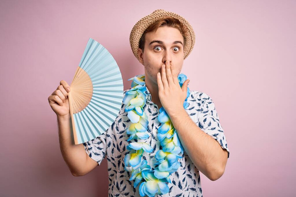 Young handsome redhead tourist man on vacation wearing hawaiian lei and hat using hand fan covering mouth with hand, shocked and afraid for mistake. Surprised expression - Фото, зображення