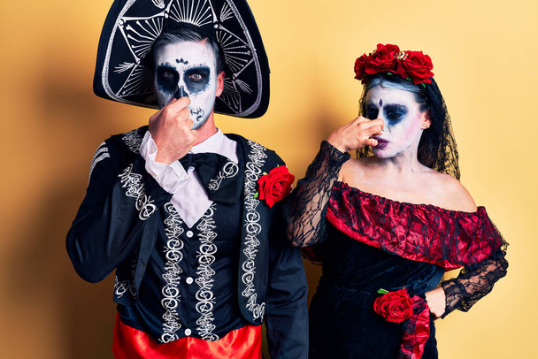 Young couple wearing mexican day of the dead costume over yellow smelling something stinky and disgusting, intolerable smell, holding breath with fingers on nose. bad smell  - Fotó, kép