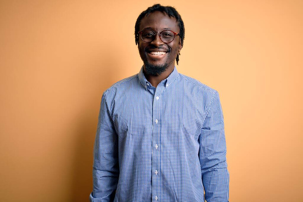 Junger gutaussehender afrikanisch-amerikanischer Mann mit Hemd und Brille vor gelbem Hintergrund und einem fröhlichen und kühlen Lächeln im Gesicht. Glücklicher Mensch. - Foto, Bild