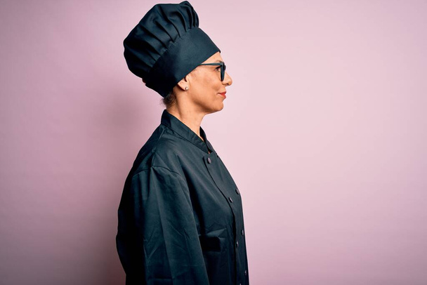 Moyen âge brunette chef femme portant uniforme de cuisinière et chapeau sur fond rose isolé regardant de côté, pose de profil relax avec visage naturel et sourire confiant. - Photo, image