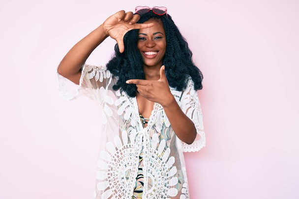 Mooie Afrikaanse vrouw in zomerjurk en zonnebril lachend lijstje makend met handen en vingers met vrolijk gezicht. creativiteit en fotografie.  - Foto, afbeelding