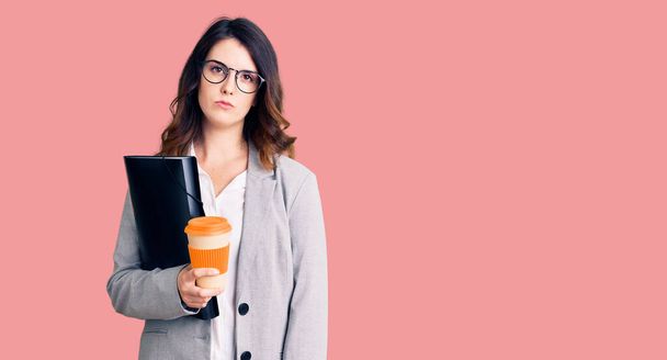 Krásná mladá brunetka žena drží obchodní složku a pití kávy myšlení postoj a střízlivý výraz hledá sebevědomí  - Fotografie, Obrázek