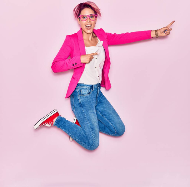 Νεαρή όμορφη επιχειρηματίας με ροζ κοντά μαλλιά φορώντας κομψά ρούχα και γυαλιά χαμογελώντας ευτυχισμένη. Άλμα με χαμόγελο στο πρόσωπο δείχνει προς την πλευρά με τα δάχτυλα πάνω από απομονωμένο φόντο - Φωτογραφία, εικόνα