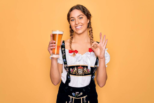 Schöne blonde deutsche Frau mit blauen Augen trägt Oktoberfestkleid, trinkt ein Glas Bier und tut ok Zeichen mit den Fingern, hervorragendes Symbol - Foto, Bild