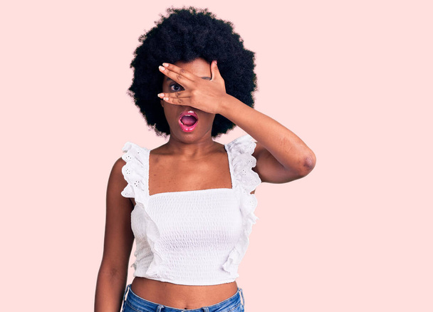 jonge afrikaanse amerikaanse vrouw dragen casual kleding gluren in shock bedekking gezicht en ogen met de hand, kijken door vingers met beschaamde uitdrukking.  - Foto, afbeelding