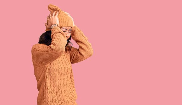 Nuori kaunis sekarotuinen nainen yllään villapaita ja talvihattu kärsivät päänsärky epätoivoinen ja stressaantunut, koska kipu ja migreeni. kädet pään päällä.  - Valokuva, kuva