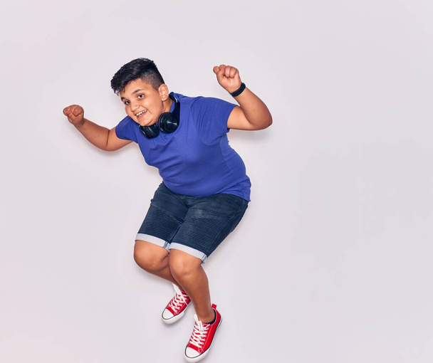Adorable niño hispano sonriendo feliz. Saltar con sonrisa en la cara escuchando música usando auriculares sobre un fondo blanco aislado - Foto, imagen