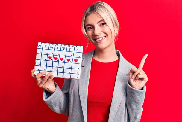 Красивая блондинка, контролирующая период менструации календаря на красном фоне улыбаясь счастливо указывая рукой и пальцем в сторону - Фото, изображение