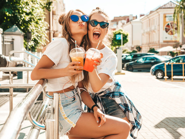 Two young beautiful smiling hipster women in trendy summer clothes. Carefree women posing outdoors.Positive models holding and drinking fresh cocktail smoothie drink in plastic cup with straw - Foto, afbeelding