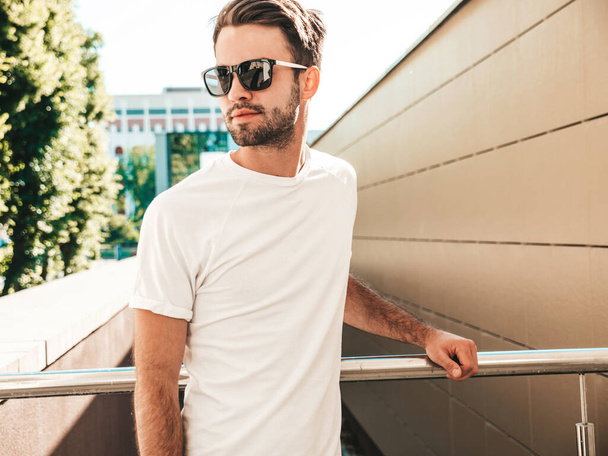 Portrait of handsome confident stylish hipster lambersexual model.Man dressed in white T-shirt. Fashion male posing on the street background in sunglasses outdoors - Foto, imagen