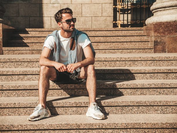 Portrait of handsome confident stylish hipster lambersexual model.Man dressed in white T-shirt. Fashion male sitting at the stairs on the street background in sunglasses - Фото, зображення