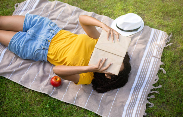 Blick auf Millennial Black Girl, das Buch auf Picknickdecke draußen liest - Foto, Bild