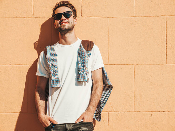 Portrait of handsome smiling stylish hipster lambersexual model.Man dressed in white T-shirt. Fashion male posing on the street background near wall in sunglasses outdoors - Φωτογραφία, εικόνα
