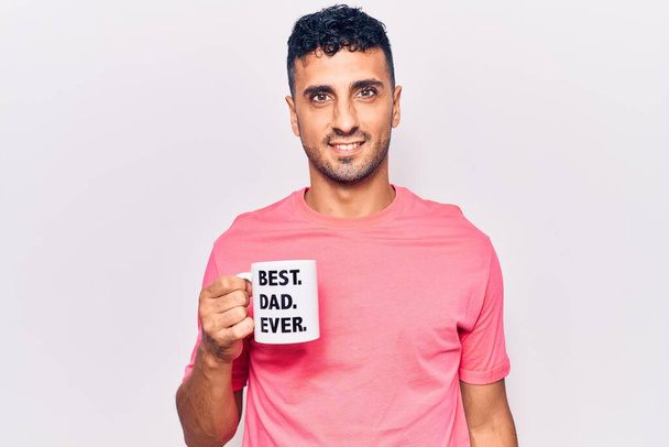 Young hispanic man drinking mug of coffe with best dad ever message looking positive and happy standing and smiling with a confident smile showing teeth  - Photo, Image
