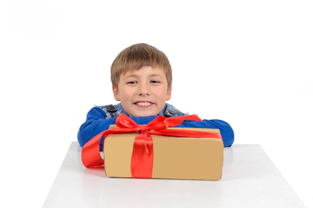 o menino está sentado na mesa, na frente dele é um presente em embalagens de papel com uma fita vermelha
 - Foto, Imagem