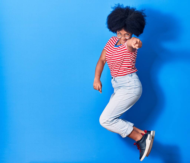 若い美しいアフリカ系アメリカ人女性のカジュアルな服や眼鏡を着て幸せな笑顔。孤立した青の背景の上に笑顔でジャンプ - 写真・画像