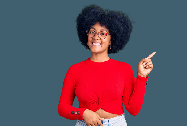 Junge Afroamerikanerin trägt lässige Kleidung und Brille mit einem breiten Lächeln im Gesicht und zeigt mit dem Finger zur Seite in die Kamera.  - Foto, Bild