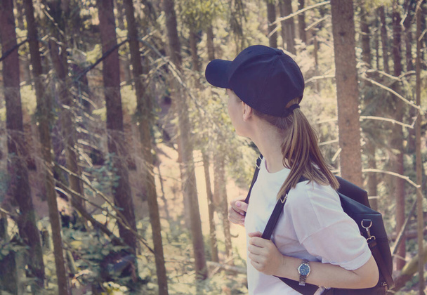 Active young female tourist with a black backpack and a cap in a coniferous forest on a Sunny day side view.Hiking.Woman tourism.individual adventures - Photo, Image