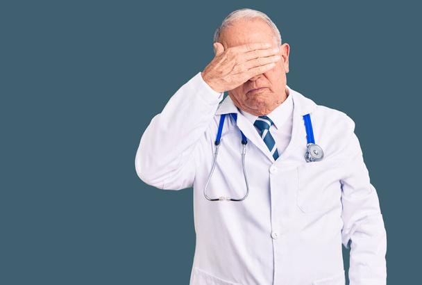 Starszy przystojny siwowłosy mężczyzna w płaszczu lekarskim i stetoskopie zasłaniający dłońmi oczy, wyglądający poważnie i smutno. Koncepcja niewidoczności, ukrywania i odrzucania  - Zdjęcie, obraz