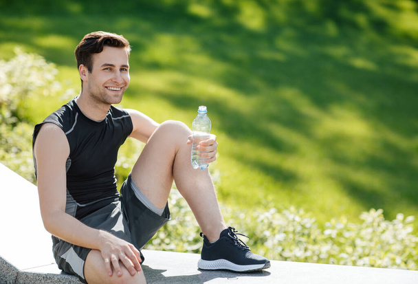 Salud y deporte. Hombre sosteniendo una botella de agua y sentado en el parque
 - Foto, imagen