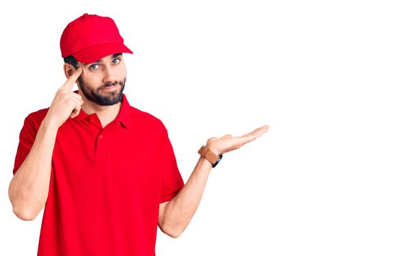Young handsome man with beard wearing delivery uniform confused and annoyed with open palm showing copy space and pointing finger to forehead. think about it.  - Photo, Image