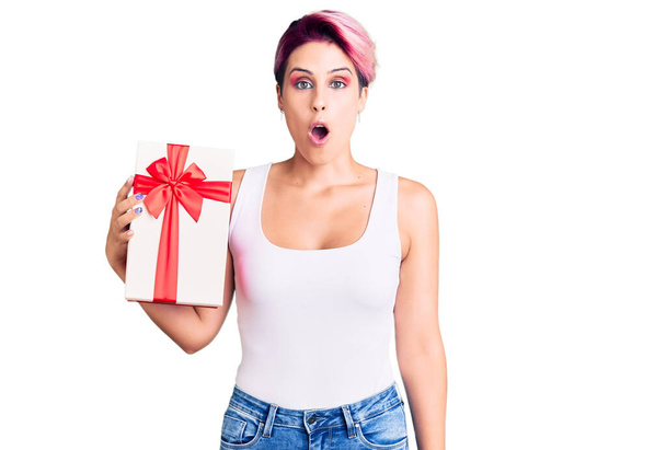 Junge schöne Frau mit rosa Haaren hält Geschenk verängstigt und erstaunt mit offenem Mund für Überraschung, ungläubiges Gesicht  - Foto, Bild