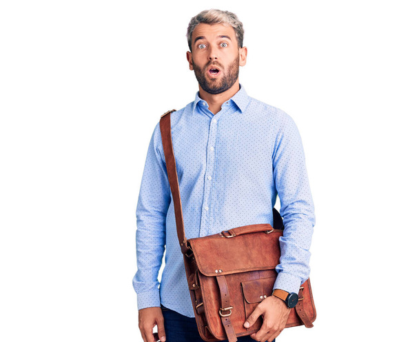 Young handsome blond man wearing elegant shirt holding briefcase scared and amazed with open mouth for surprise, disbelief face  - Foto, Imagen