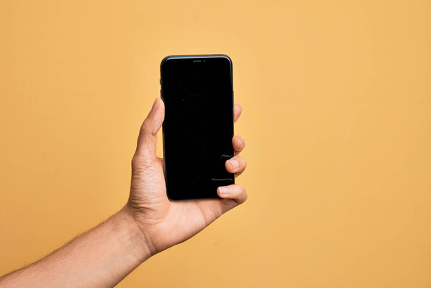 Hand van de Kaukasische jongeman die smartphone toont scherm over geïsoleerde gele achtergrond - Foto, afbeelding