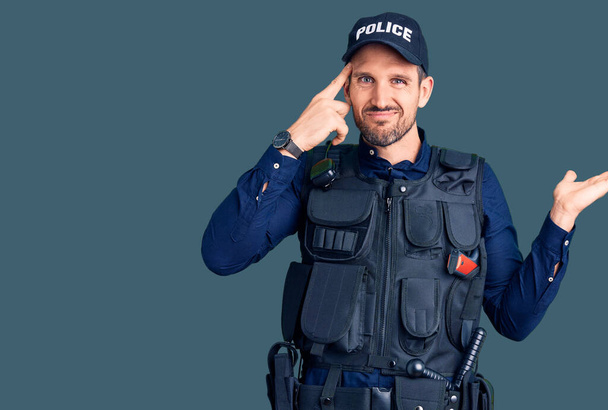 Jonge knappe man draagt politie-uniform verward en geïrriteerd met open palm tonen kopieerruimte en wijzende vinger naar het voorhoofd. Denk er eens over na..  - Foto, afbeelding