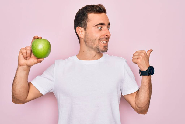 Homem bonito jovem com olhos azuis comendo maçã verde saudável sobre fundo rosa isolado apontando e mostrando com o polegar até o lado com o rosto feliz sorrindo
 - Foto, Imagem
