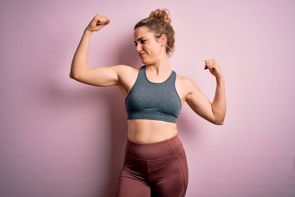 Jonge mooie blonde sportvrouw doet sport dragen sportkleding over roze achtergrond tonen armen spieren glimlachen trots. Fitness concept. - Foto, afbeelding