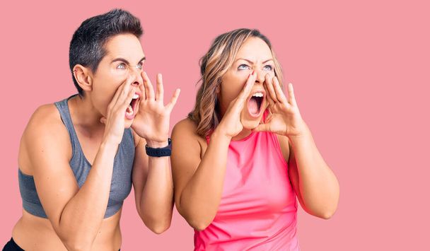 Couple de femmes portant des vêtements de sport criant en colère à haute voix avec les mains sur la bouche  - Photo, image