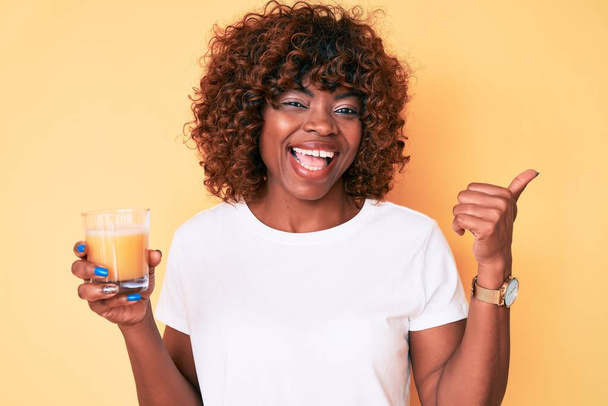 Nuori afrikkalainen amerikkalainen nainen juo lasillinen appelsiinimehua osoittaa peukalolla sivulle hymyillen onnellinen suu auki  - Valokuva, kuva