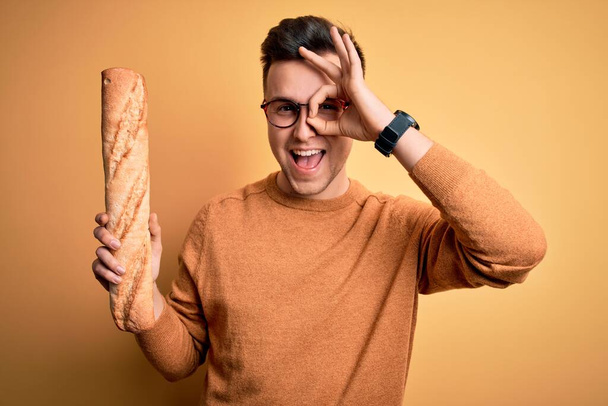 Νεαρός όμορφος καυκάσιος άνδρας που φοράει γυαλιά κρατώντας μια φρέσκια μπαγκέτα ψωμί με χαρούμενο πρόσωπο χαμογελώντας κάνοντας OK σημάδι με το χέρι στο μάτι κοιτάζοντας μέσα από τα δάχτυλα - Φωτογραφία, εικόνα