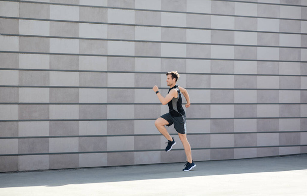 Training and running in city in morning. Millennial man in sportswear and fitness tracker runs and jumps - Φωτογραφία, εικόνα