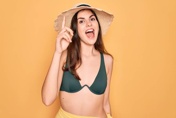 Young beautiful girl wearing swimwear bikini and summer sun hat over yellow background pointing finger up with successful idea. Exited and happy. Number one. - Valokuva, kuva