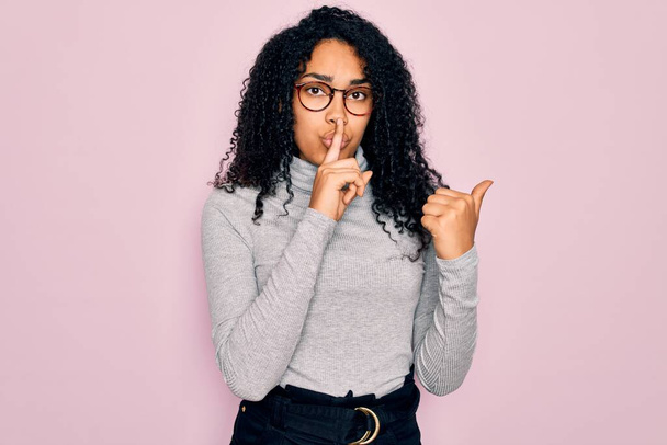 Mujer afroamericana joven con jersey de cuello alto y gafas sobre fondo rosa pidiendo estar callada con el dedo en los labios apuntando con la mano hacia un lado. Silencio y concepto secreto
. - Foto, imagen