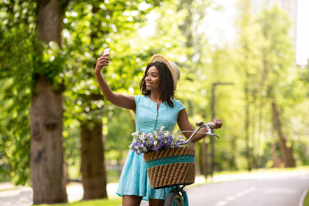 Υπέροχο μαύρο κορίτσι λαμβάνοντας selfie για βόλτα με το ποδήλατο στο ηλιόλουστο καλοκαιρινό πάρκο - Φωτογραφία, εικόνα